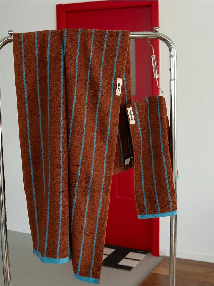 A brown terry cloth towel with vertical turquoise blue stripes and matching blue trim hangs on a silver metal rack in front of a red door.