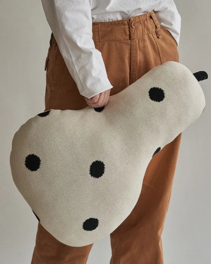 A person wearing a white shirt and brown pants holds a large pear-shaped knit cushion with black polka dots against a plain light gray background.