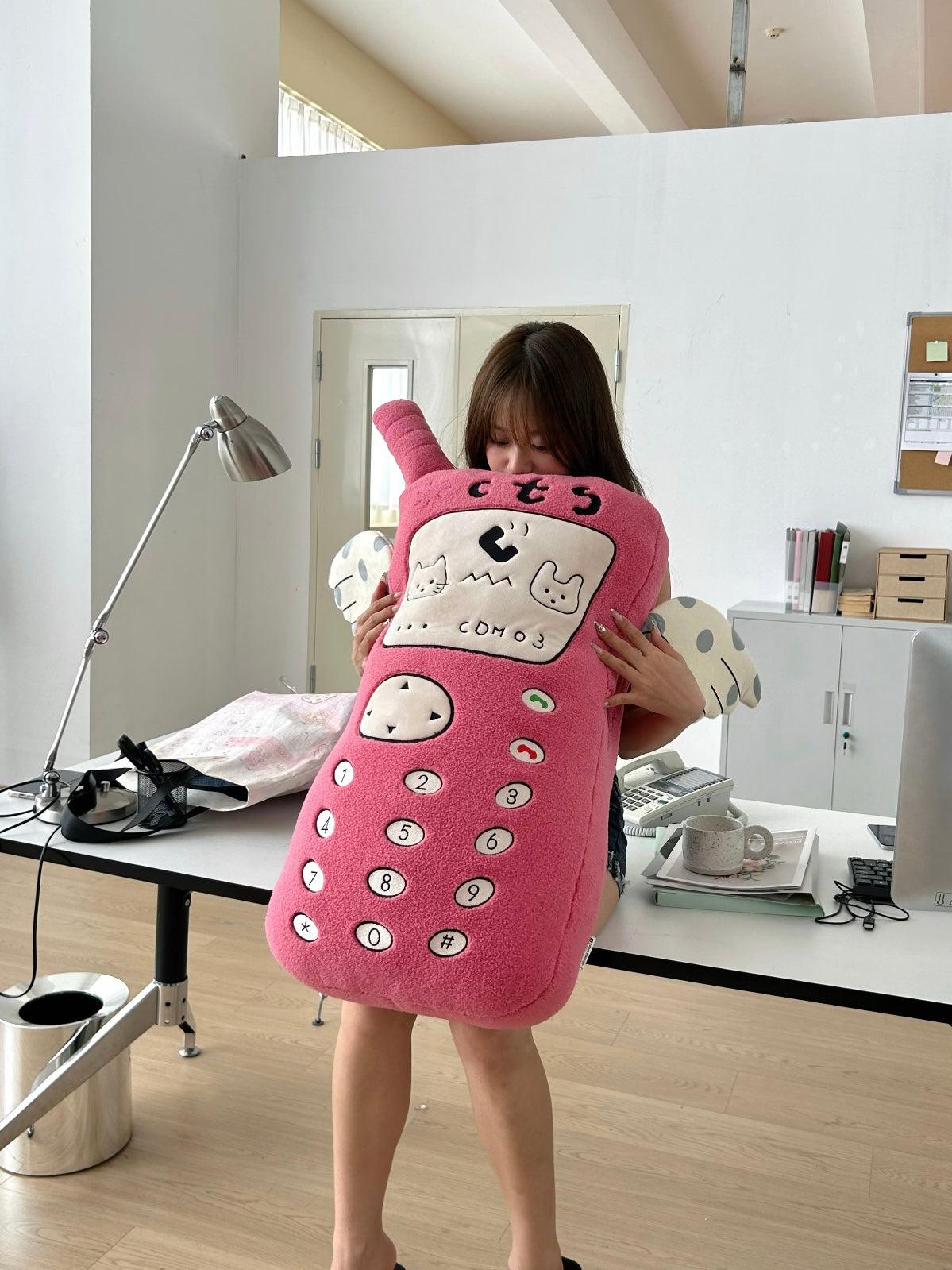 A person holds a pink telephone plush upright, with the keypad facing forward and spotted arms at the sides.