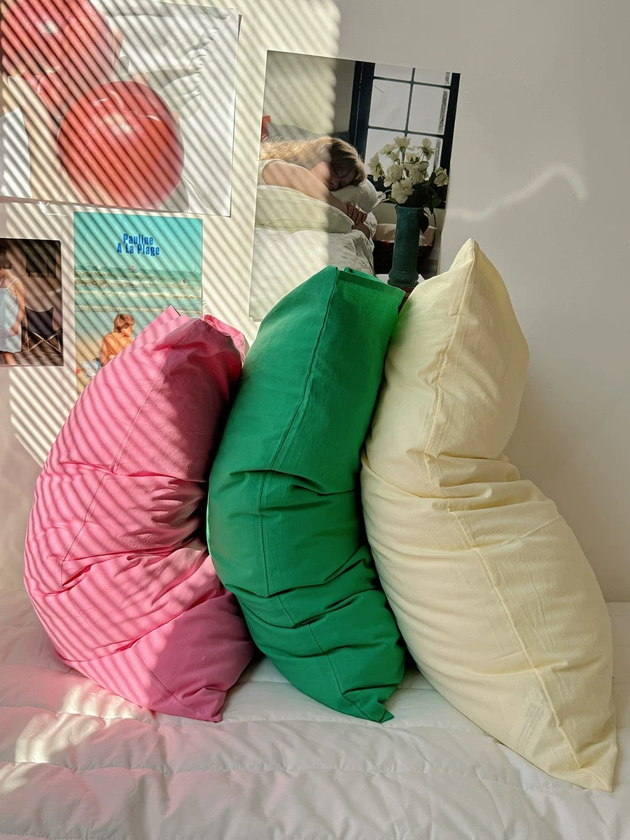 Three colorful pillows stacked on a bed: a bright pink pillow on top, a pale yellow pillow in the middle, and a green pillow at the bottom, set against a white wall with decorative posters and window light.