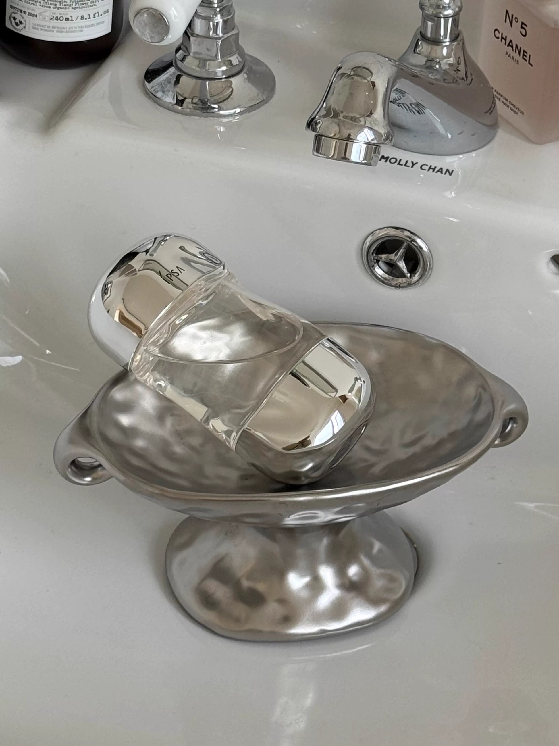 A clear glass perfume bottle with a silver cap resting on a hammered silver metal soap dish placed on a white ceramic bathroom sink. A Chanel bottle is partially visible in the background.