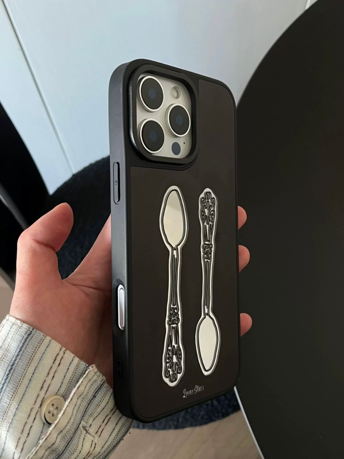 A vintage phone case is held upright, displaying two ornate spoons in opposite directions on a black back.