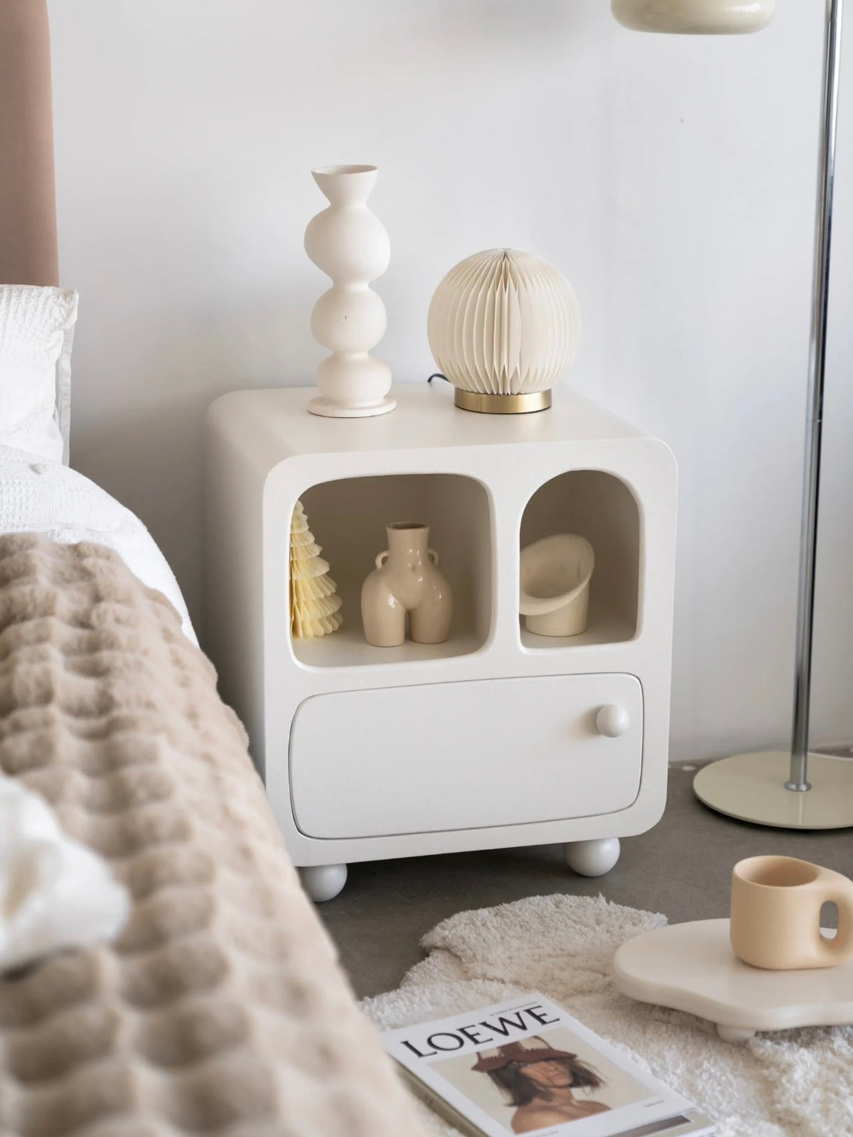 A white arched nightstand shown closer has small vases inside its two openings and round feet below.