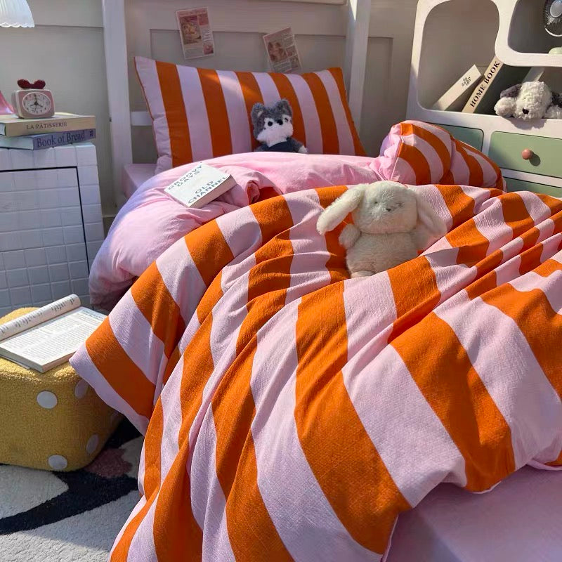 A cozy bedroom scene showing a bed made with a pink and orange striped bedding set, including a striped pillowcase, matching striped pillow, pink duvet cover, and striped comforter. A white bunny plush toy rests on the bedding, with a white nightstand, shelves, and other bedroom items in the background.