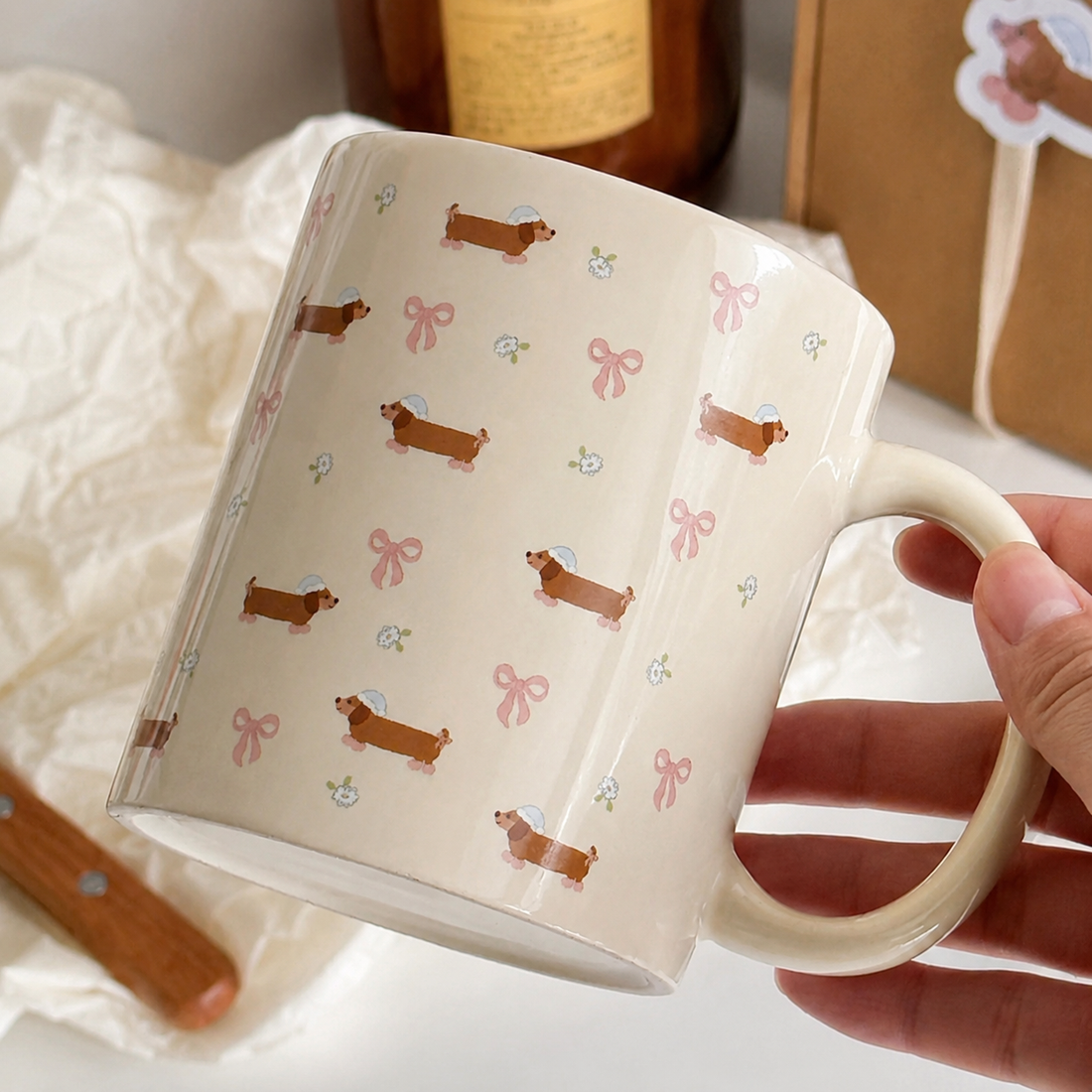 A cream-colored ceramic mug with a repeating pattern of brown dachshunds wearing pink bows, nestled inside a woven wire basket lined with white tissue paper. A matching dachshund sticker is placed on the front of the mug.