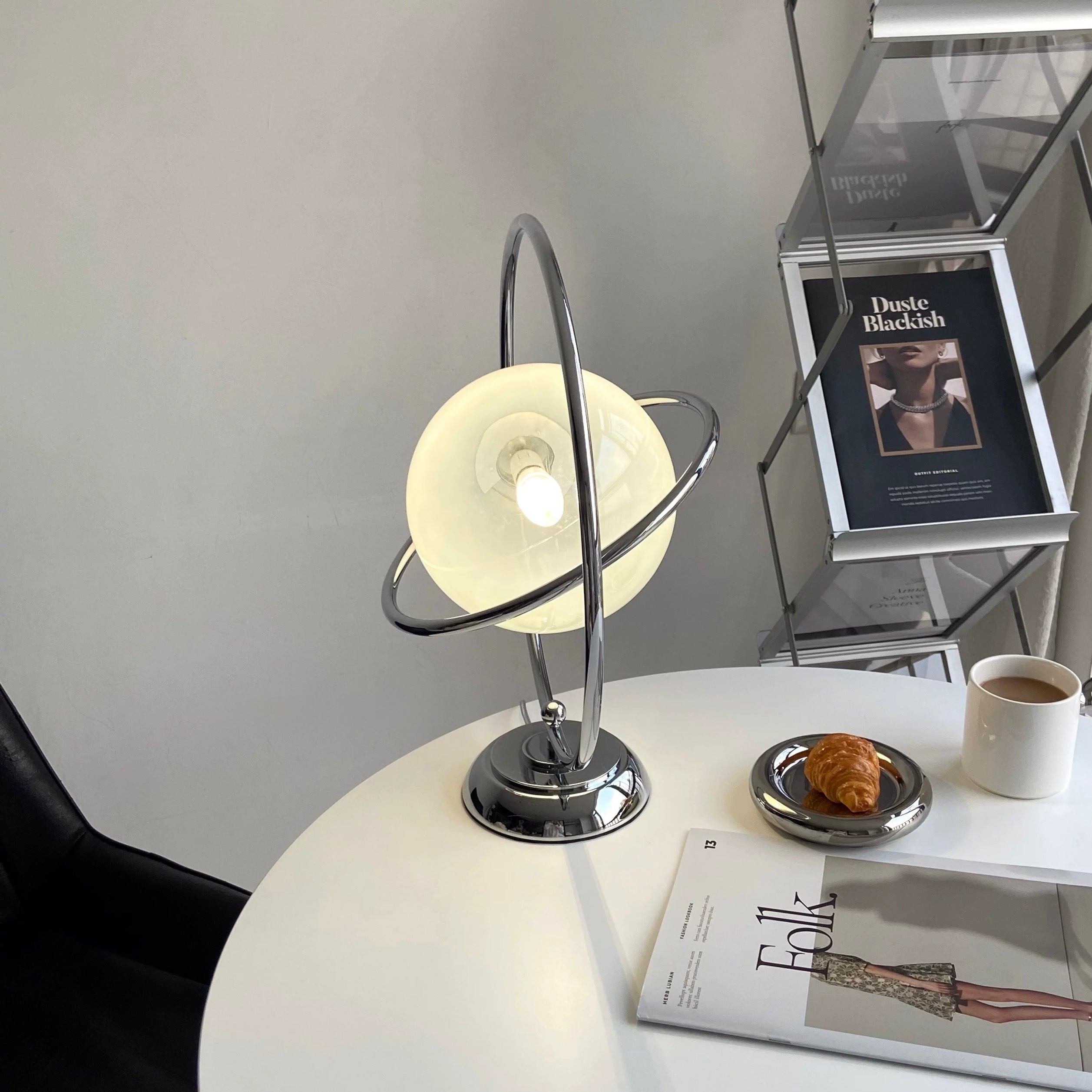 Modern table lamp on a round white table with a cup, pastry, and magazine in a minimalistic room.