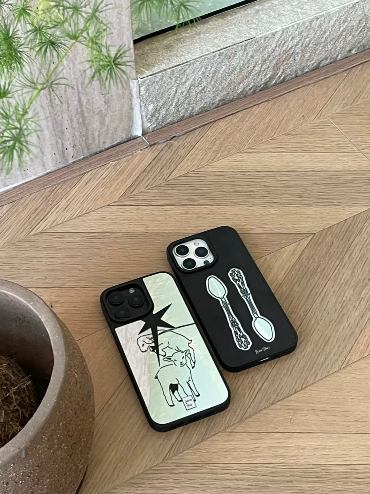 A Y2K phone case with silver lamb and star artwork lies beside a black case printed with two vintage spoons.