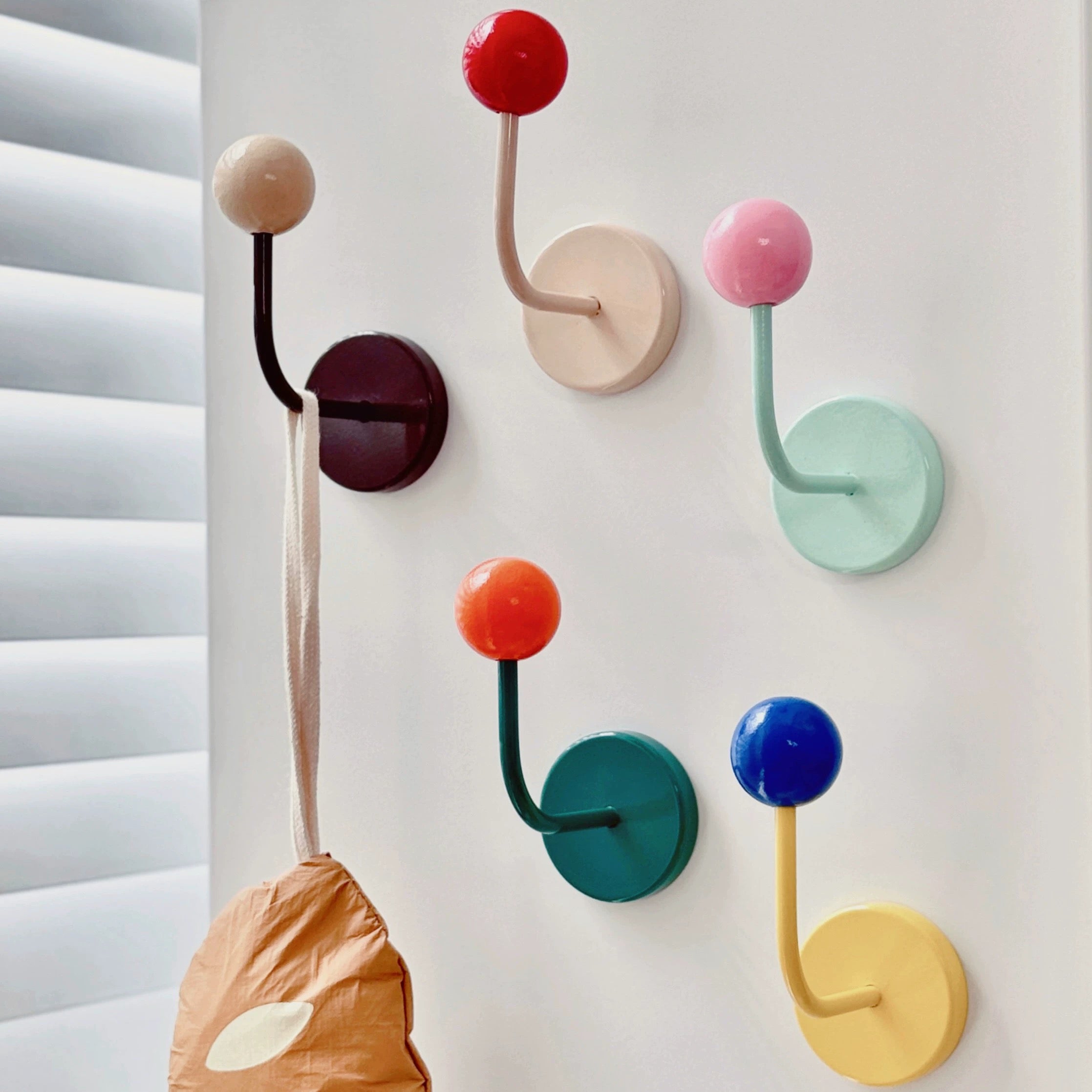 Six wall-mounted coat hooks in assorted pastel and bright colors, arranged on a white wall next to window blinds. A tan paper bag hangs from one hook on the left.