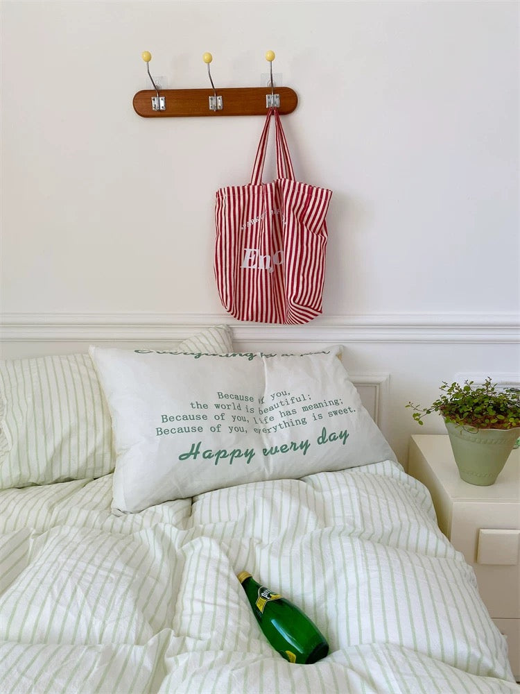 Red and white striped tote bag hanging on a wooden hook above a bed with a pillow and bottle.